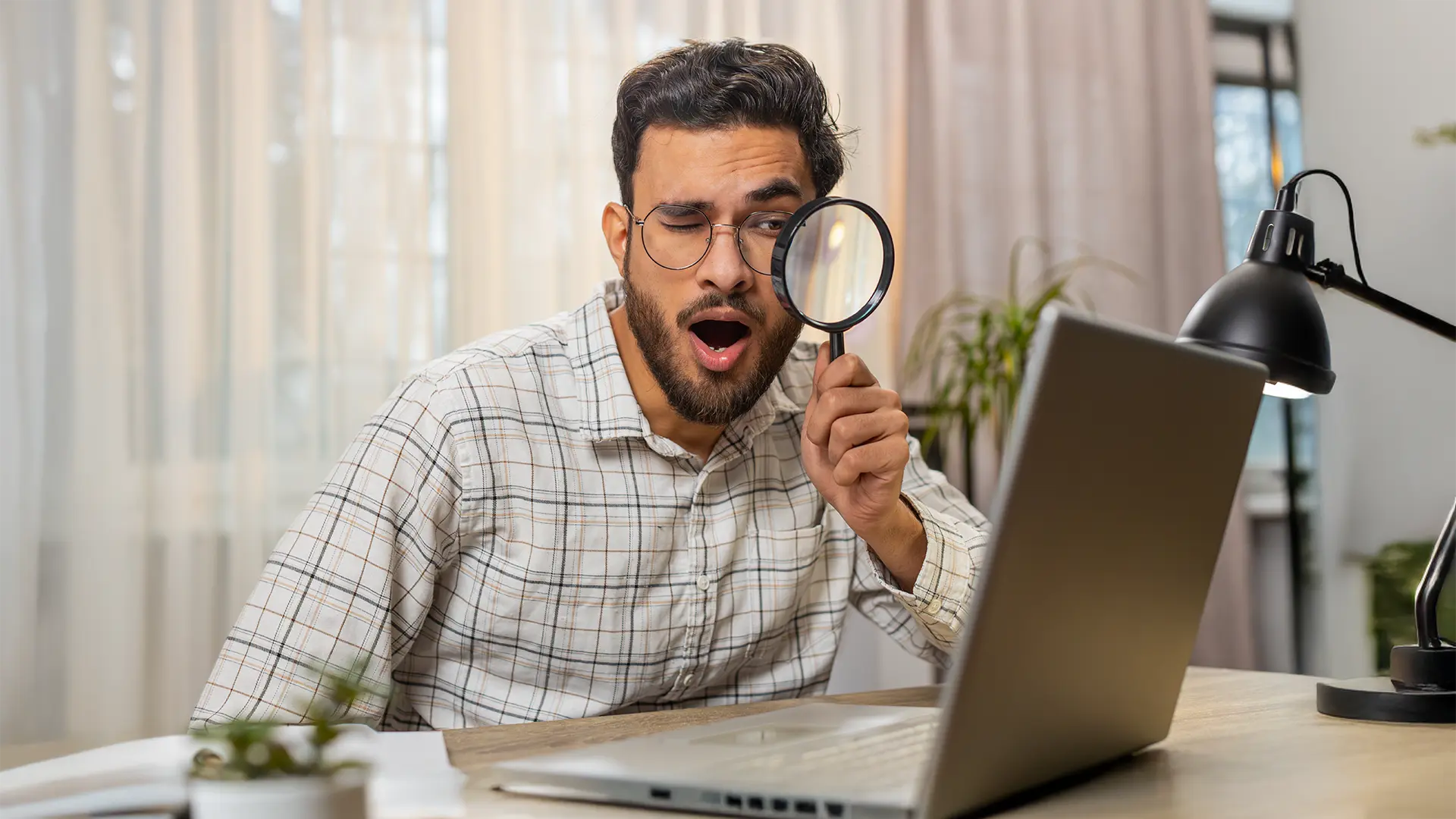 A middle aged, brunette man is sitting at a desk, looking through a magnifying glass at his laptop. He has an exaggerated surprised expression on his face.
