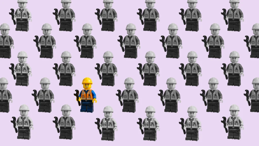 A grid of identical grey Lego construction worker figurines, with a single colourful yellow-and-orange Lego worker standing out in the centre of the crowd. Image of a hard hat hanging on a hat stand.